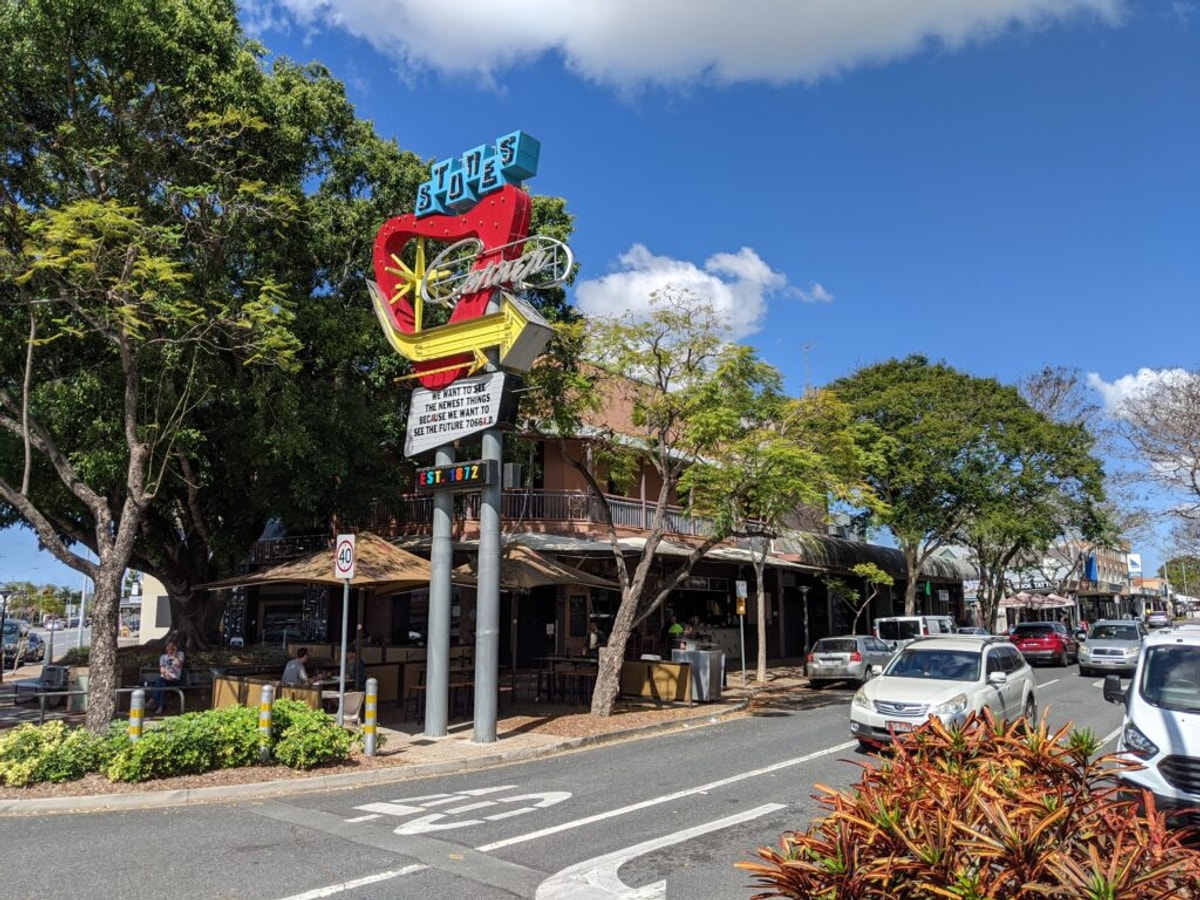 A brand new, but retro looking sign stands above the entryway to Stones Corner. People are dining on the footpath, and a couple of cars roll past.