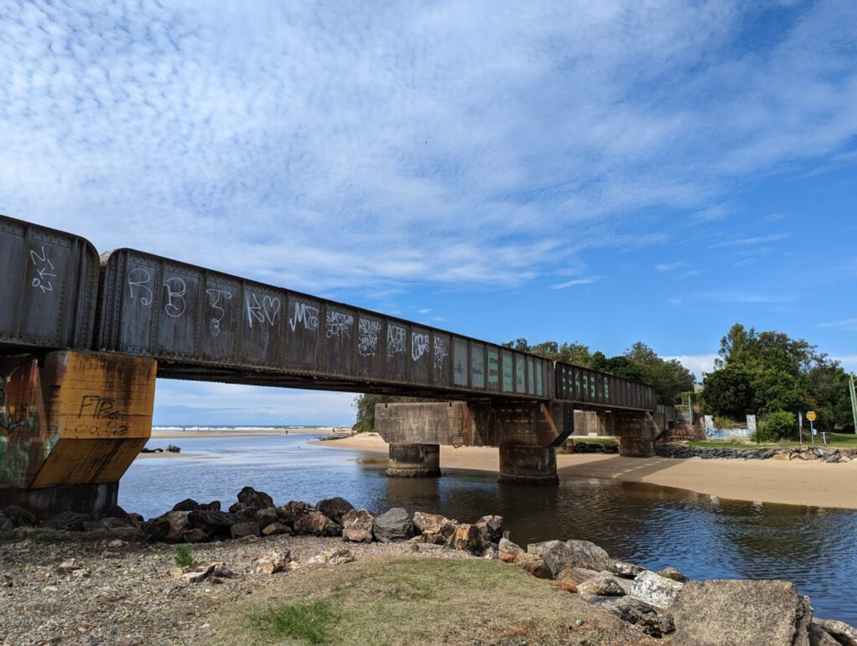 A concrete and steel bridge spans a tea stained creek, the beach in the distance.