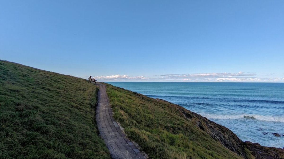 A man sits by himself at a bench overlooking the ocean, on the side of a steep hill, with a paved path trailing into the distance.