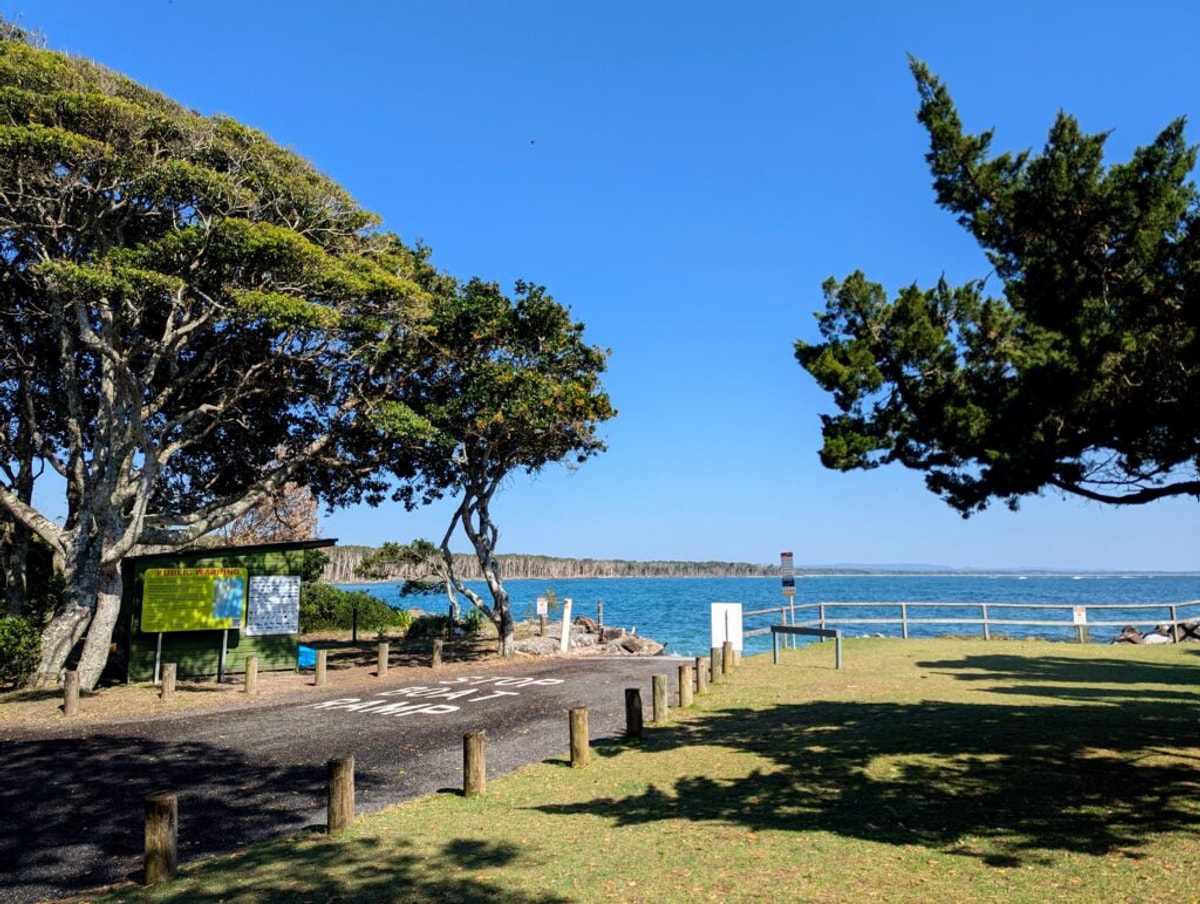 A road leads to a boat ramp in a cerulean sea, a giant tree shades a green wooden beach shack.