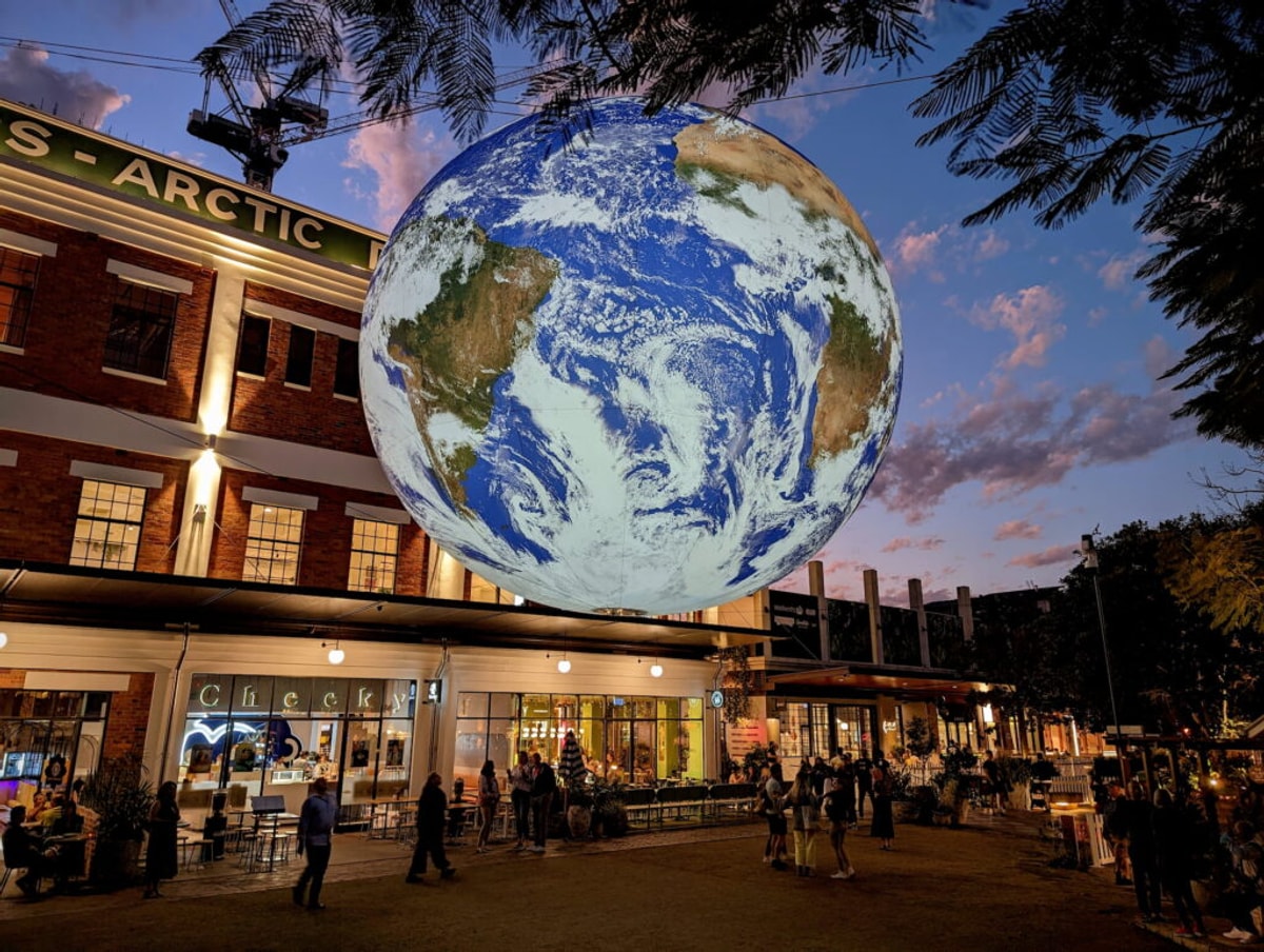 A twilight shot of a grassy square, an old gentrified factory building in the background, and a giant illuminated planet earth suspended in the air. South America and Africa are visible. There are people admiring underneath.