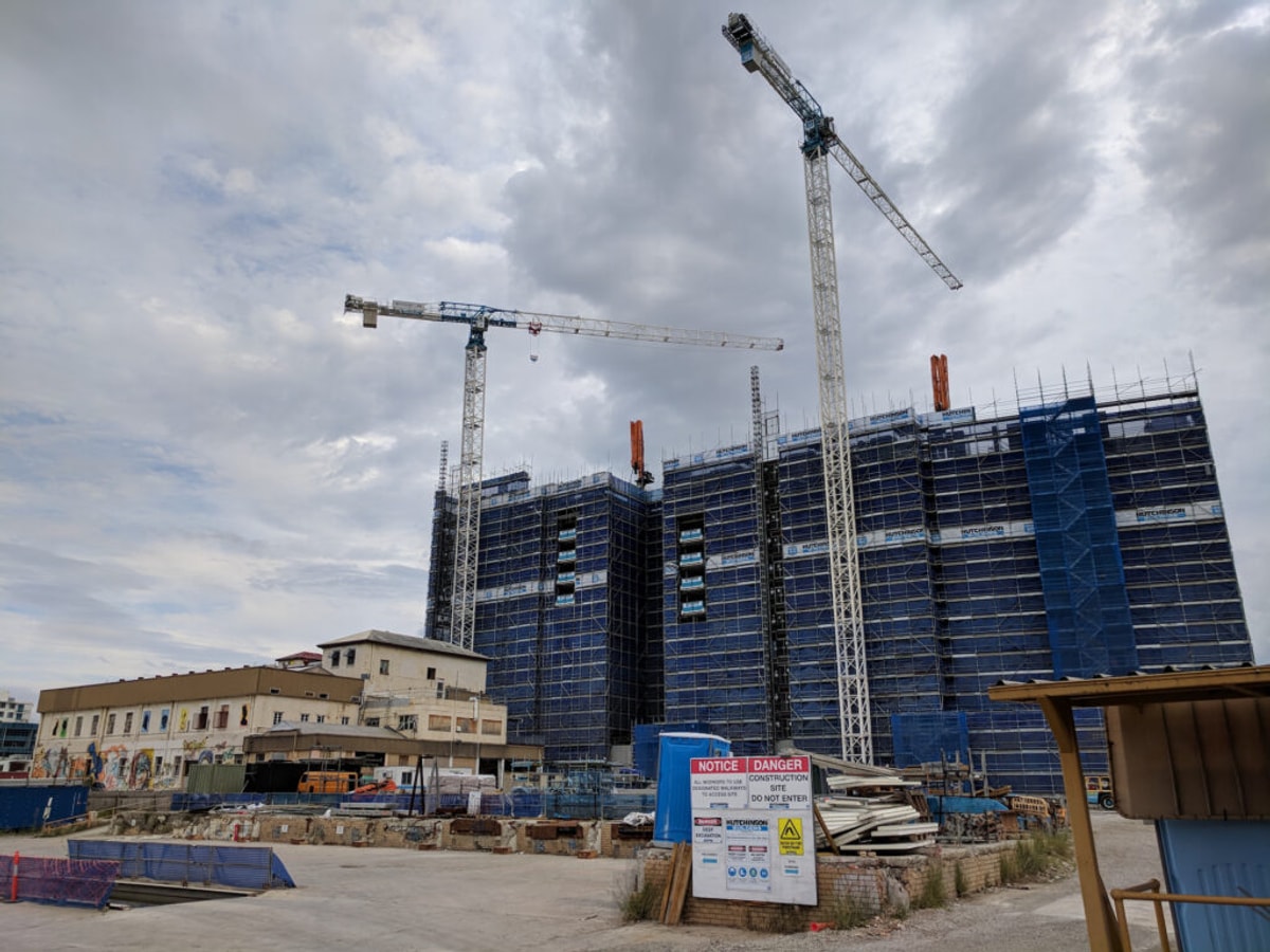 A concrete building site. A two story historic factory sits in front of three massive, blocky towers under construction.